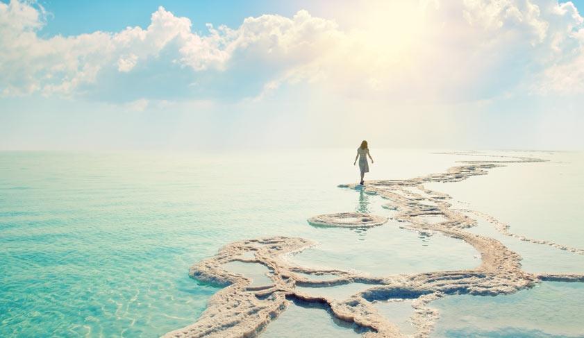 Silhouette of young woman walking on Dead Sea salt shore at sunrise towards the sun.